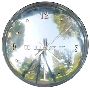 May include: A round, silver-colored clock with a reflective face. The clock features gold-colored hands and hour markers, with the numbers 12, 3, 6, and 9 clearly visible. The clock's face reflects a blurry outdoor scene.