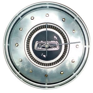 May include: A round clock made from a silver hubcap. The clock face is black with a silver emblem and black clock hands. The hubcap has silver rivets around the edge.