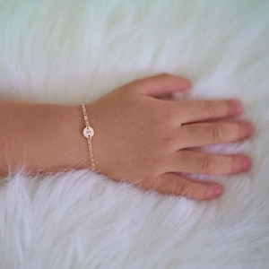 May include: A delicate gold-colored bracelet with a small, round charm featuring the letter "E". The bracelet is on a chain and is shown on a person's wrist. The background is a soft, white, fluffy surface.