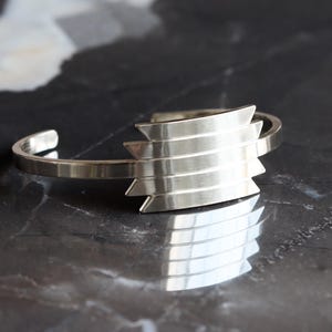 May include: A silver cuff bracelet with a geometric design. The bracelet features a series of horizontal bars with pointed edges, creating a layered effect. The metal has a polished finish, reflecting light. The bracelet is displayed on a dark, reflective surface.