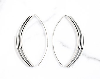 May include: A pair of silver-toned, open-ended hoop earrings. Each earring has a curved, elongated shape with a series of parallel lines and a small rectangular accent near the center. The earrings are displayed against a white marble background.