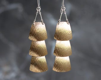 Puede incluir: Un par de pendientes dorados en capas. Cada pendiente presenta tres colgantes texturizados en forma de trapecio suspendidos de un gancho plateado. Los pendientes cuelgan de un alambre plateado, con un fondo gris oscuro borroso.