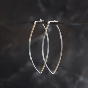 May include: A pair of silver-toned hoop earrings. The earrings have a slender, elongated oval shape, with a pointed tip at the bottom and a hook closure at the top. The earrings are suspended against a dark background.