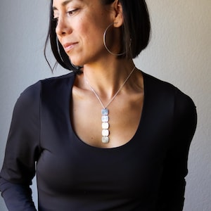 May include: A silver necklace with a series of square pendants hangs on a black top. The woman is wearing large silver hoop earrings. The background is a solid light gray.