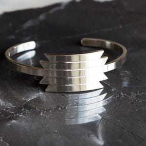May include: A silver cuff bracelet with a geometric design. The bracelet features a series of horizontal bars that form a stepped pattern in the center. The metal has a polished finish, reflecting light. The bracelet is displayed on a dark, textured surface.