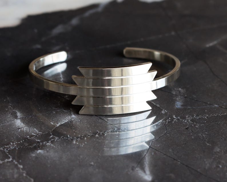 May include: A silver cuff bracelet with a geometric design. The bracelet features a series of horizontal bars that form a stepped pattern in the center. The metal has a polished finish, reflecting light. The bracelet is displayed on a dark, textured surface.