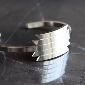 May include: A silver cuff bracelet with a geometric design. The bracelet features a series of layered, pointed shapes on the front, creating a modern and stylish look. The polished metal reflects light, adding to its appeal. This accessory is suitable for everyday wear.