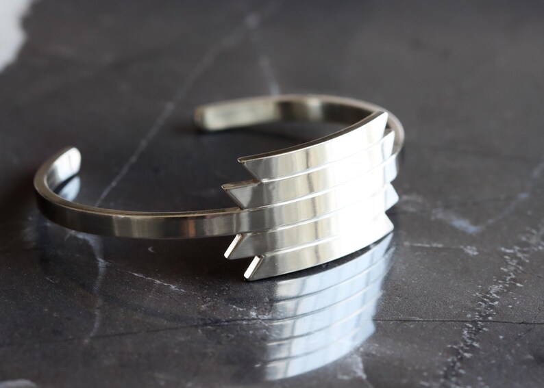 May include: A silver cuff bracelet with a geometric design. The bracelet features a series of layered, pointed shapes on the front, creating a modern and stylish look. The polished metal reflects light, adding to its appeal. This accessory is suitable for everyday wear.