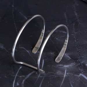 Puede incluir: Una pulsera de pu&ntilde;o plateada con un dise&ntilde;o minimalista. La pulsera presenta dos bandas curvas y paralelas conectadas por una sola barra. Los extremos abiertos de la pulsera est&aacute;n ligeramente aplanados. La joya se exhibe sobre una superficie oscura y veteada.