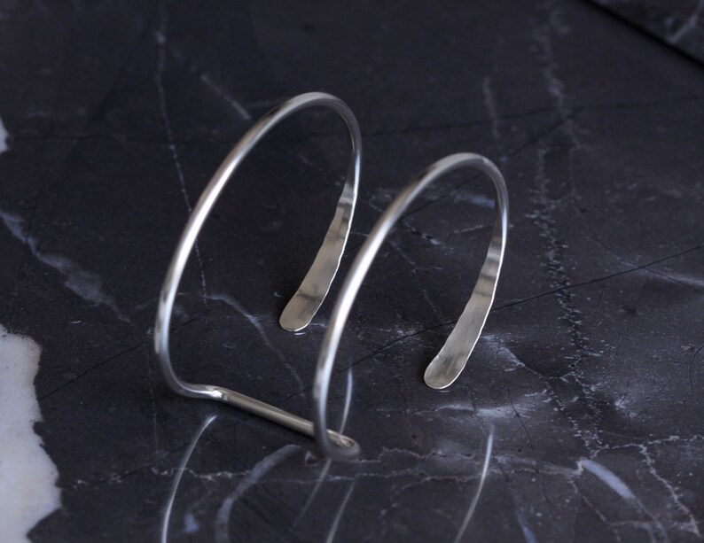 Puede incluir: Una pulsera de pu&ntilde;o plateada con un dise&ntilde;o minimalista. La pulsera presenta dos bandas curvas y paralelas conectadas por una sola barra. Los extremos abiertos de la pulsera est&aacute;n ligeramente aplanados. La joya se exhibe sobre una superficie oscura y veteada.
