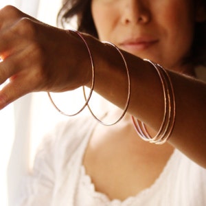 May include: Several thin, rose gold bangles are displayed on a person's arm. The bangles are simple, circular, and have a hammered texture. The person is wearing a white top. The background is softly lit.