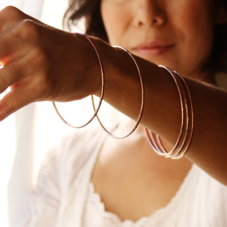 May include: Several thin, rose gold bangles are displayed on a person's arm. The bangles are simple, circular, and have a hammered texture. The person is wearing a white top. The background is softly lit.