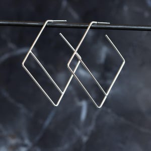 Op de afbeelding: Een paar zilveren geometrische oorbellen. Elke oorbel heeft een vierkante vorm met een diagonale binnenlijn. De oorbellen hangen tegen een donkere achtergrond. Ze zijn gemaakt van glanzend metaal.