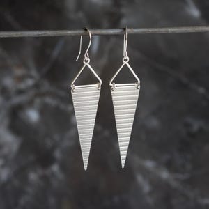Puede incluir: Un par de pendientes plateados. Cada pendiente presenta un colgante triangular con líneas horizontales grabadas, suspendido de un pequeño triángulo y un gancho. Fondo oscuro y borroso.