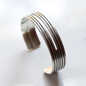 Puede incluir: Una pulsera de plata con un diseño moderno. La pulsera presenta múltiples bandas paralelas, creando un efecto de capas. El metal pulido refleja la luz, resaltando el estilo elegante y minimalista. La pulsera mide aproximadamente 6,35 cm de ancho.