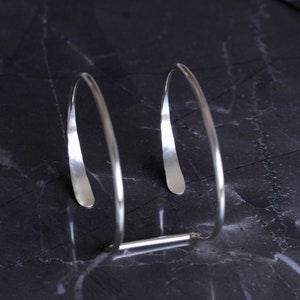 Puede incluir: Una pulsera de plata minimalista con un dise&ntilde;o abierto &uacute;nico. La pulsera presenta dos elementos curvos y alargados conectados por una peque&ntilde;a barra. El metal pulido refleja la luz, creando una est&eacute;tica elegante y moderna. La pulsera se muestra sobre una superficie oscura y veteada.