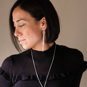Peut inclure: Une femme portant un pull noir et des bijoux en argent. Les bijoux comprennent de longues boucles d'oreilles pendantes et un collier. Les boucles d'oreilles ont un design lin&eacute;aire, et le collier est une simple cha&icirc;ne. La femme a les cheveux fonc&eacute;s et regarde vers le bas.