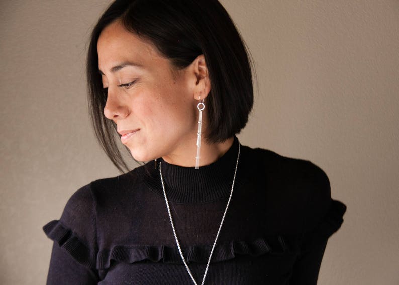 Peut inclure: Une femme portant un pull noir et des bijoux en argent. Les bijoux comprennent de longues boucles d'oreilles pendantes et un collier. Les boucles d'oreilles ont un design lin&eacute;aire, et le collier est une simple cha&icirc;ne. La femme a les cheveux fonc&eacute;s et regarde vers le bas.