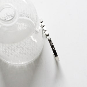 May include: A silver-toned bracelet with a series of small, evenly spaced, circular accents. The bracelet is positioned next to a clear glass object with a geometric pattern, all set against a white background. The bracelet's design is minimalist and modern.