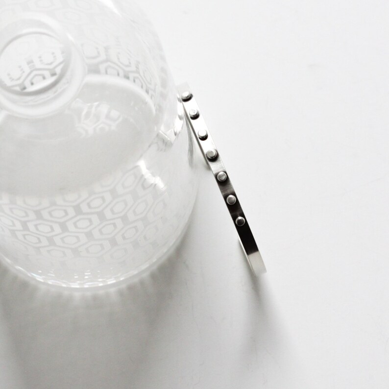 May include: A silver-toned bracelet with a series of small, evenly spaced, circular accents. The bracelet is positioned next to a clear glass object with a geometric pattern, all set against a white background. The bracelet's design is minimalist and modern.