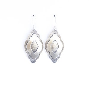 Pode incluir: Um par de brincos pendurados prateados. Cada brinco apresenta um design em camadas com uma camada externa texturizada e uma camada interna lisa e vazada. Os brincos penduram-se em ganchos prateados simples sobre um fundo branco.