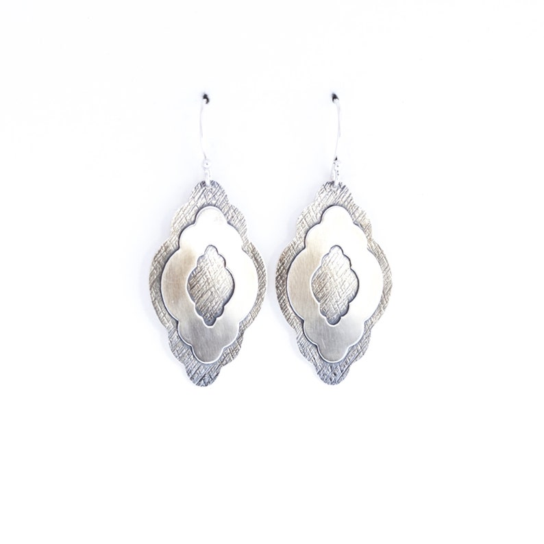 Pode incluir: Um par de brincos pendurados prateados. Cada brinco apresenta um design em camadas com uma camada externa texturizada e uma camada interna lisa e vazada. Os brincos penduram-se em ganchos prateados simples sobre um fundo branco.