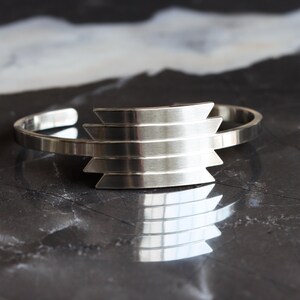 May include: A silver cuff bracelet with a geometric design. The bracelet features a series of horizontal, stepped bars in the center. The open-ended design allows for easy wear. The bracelet is displayed on a dark, reflective surface.