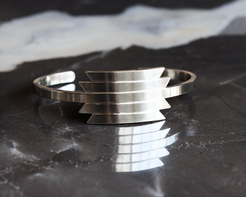 May include: A silver cuff bracelet with a geometric design. The bracelet features a series of horizontal, stepped bars in the center. The open-ended design allows for easy wear. The bracelet is displayed on a dark, reflective surface.