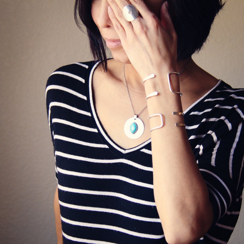Puede incluir: Juego de joyas de plata que incluye un anillo, pulseras y un collar con un colgante turquesa. Las joyas se llevan con una camiseta a rayas blancas y negras. Las pulseras son abiertas.