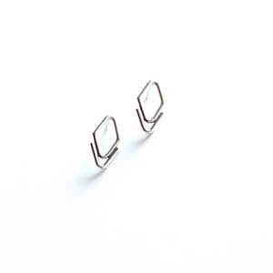 May include: Two silver-colored, geometric stud earrings. Each earring features a diamond-shaped top and a curved, angled bottom, creating a layered effect. The earrings are set against a bright white background.