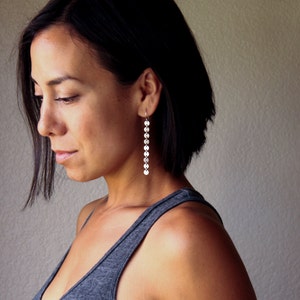 Puede incluir: Una mujer lleva pendientes largos y colgantes de plata. Los pendientes est&aacute;n compuestos por m&uacute;ltiples discos peque&ntilde;os, redondos y reflectantes que cuelgan verticalmente. La mujer lleva una camiseta sin mangas gris.