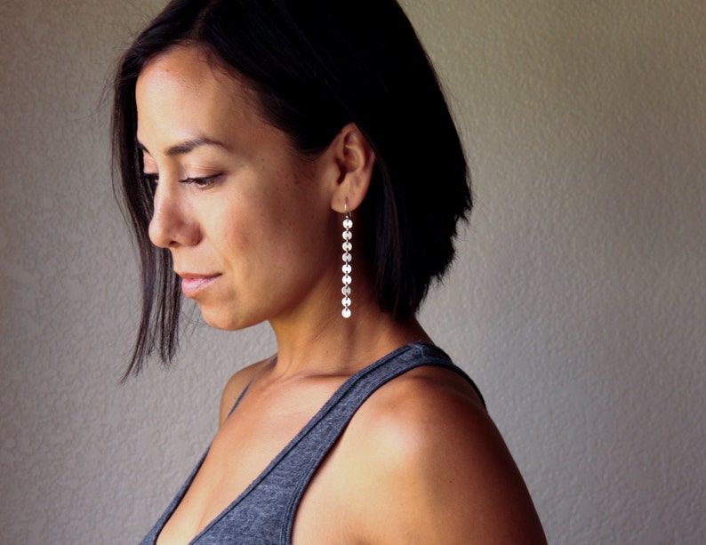 Puede incluir: Una mujer lleva pendientes largos y colgantes de plata. Los pendientes est&aacute;n compuestos por m&uacute;ltiples discos peque&ntilde;os, redondos y reflectantes que cuelgan verticalmente. La mujer lleva una camiseta sin mangas gris.