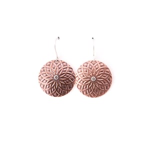 May include: A pair of copper-coloured, round earrings with a floral pattern. Each earring features a central silver-coloured stud and a decorative, geometric design. The earrings hang from silver-coloured ear wires, set against a white background.