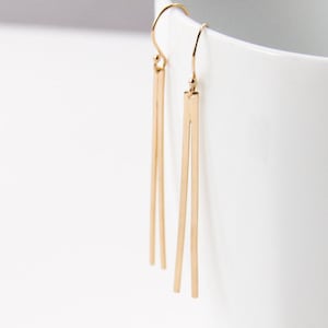 May include: Gold-colored, minimalist dangle earrings. Each earring features a simple hook and two long, rectangular bars that hang vertically. The earrings are displayed against a white background, highlighting their sleek design and metallic finish.