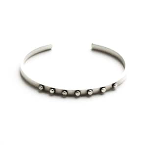 May include: A silver cuff bracelet with seven small, evenly spaced, round studs. The bracelet has a simple, minimalist design and is open at the ends. The jewelry is displayed on a white background.