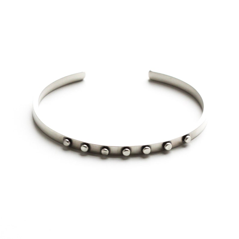 May include: A silver cuff bracelet with seven small, evenly spaced, round studs. The bracelet has a simple, minimalist design and is open at the ends. The jewelry is displayed on a white background.