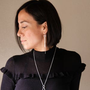 Peut inclure: Une femme porte un pull noir &agrave; col roul&eacute; et des bijoux en argent. Les bijoux comprennent de longues boucles d'oreilles pendantes et un collier d&eacute;licat avec un pendentif. Le pull a un d&eacute;tail volant&eacute; sur les &eacute;paules.