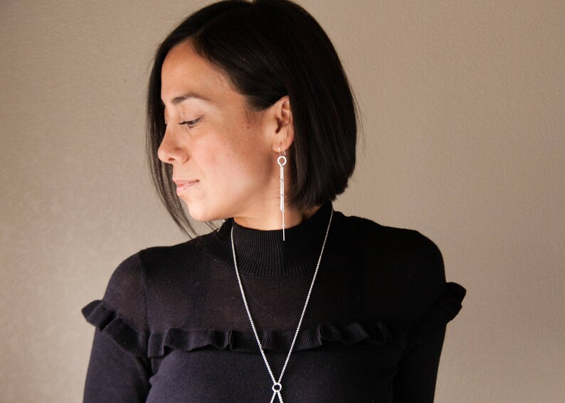 Peut inclure: Une femme porte un pull noir &agrave; col roul&eacute; et des bijoux en argent. Les bijoux comprennent de longues boucles d'oreilles pendantes et un collier d&eacute;licat avec un pendentif. Le pull a un d&eacute;tail volant&eacute; sur les &eacute;paules.