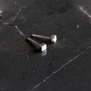 Puede incluir: Un par de pequeños pendientes de botón cuadrados de plata. Cada pendiente tiene un poste y una cabeza cuadrada. Los pendientes se muestran sobre una superficie oscura y texturizada, creando un contraste. Diseño minimalista y moderno.