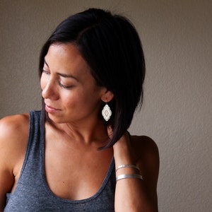 Pode incluir: Uma mulher usando uma regata cinza e pulseiras de prata. Ela est&aacute; usando um par de brincos brancos e prateados com um design geom&eacute;trico. Os brincos est&atilde;o pendurados em suas orelhas.