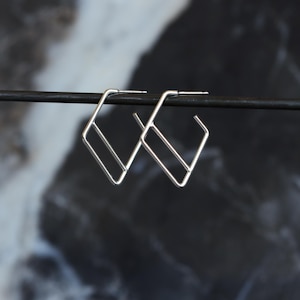 May include: A pair of geometric silver earrings. Each earring features a square shape with an interior bar, creating a modern, minimalist design. The earrings are displayed against a dark, blurred background.