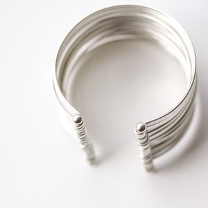 May include: A silver cuff bracelet with a layered design. The bracelet has a wide band with multiple parallel lines, creating a textured effect. The ends of the bracelet are rounded with small, decorative spheres.