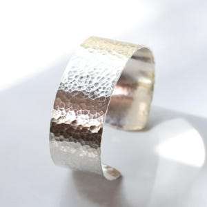 May include: A wide, hammered silver cuff bracelet. The bracelet has a textured surface with a series of small, irregular indentations. The open cuff design allows for easy wear. The jewelry is displayed on a white surface.