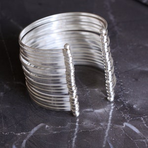 May include: A silver cuff bracelet with multiple parallel bands. The bracelet has a modern design with a series of thin, curved metal strips held together by vertical, beaded supports. The jewelry is displayed on a dark, textured surface.