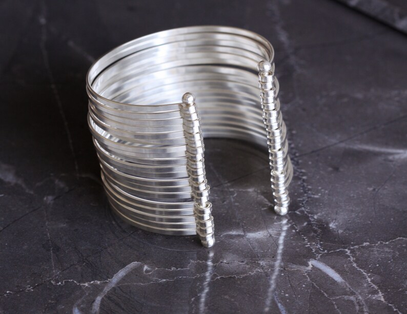 May include: A silver cuff bracelet with multiple parallel bands. The bracelet has a modern design with a series of thin, curved metal strips held together by vertical, beaded supports. The jewelry is displayed on a dark, textured surface.
