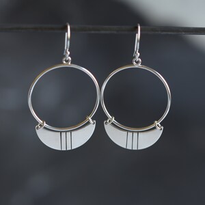 May include: A pair of silver hoop earrings. Each earring features a large, circular hoop with a half-moon shaped pendant at the bottom. The pendant has three vertical lines etched into its surface. The earrings are displayed against a dark background.