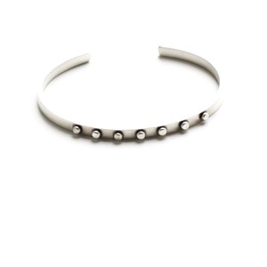 May include: A silver-colored cuff bracelet with a row of small, round, silver studs. The bracelet has an open design and is set against a white background. The jewelry is simple and modern.