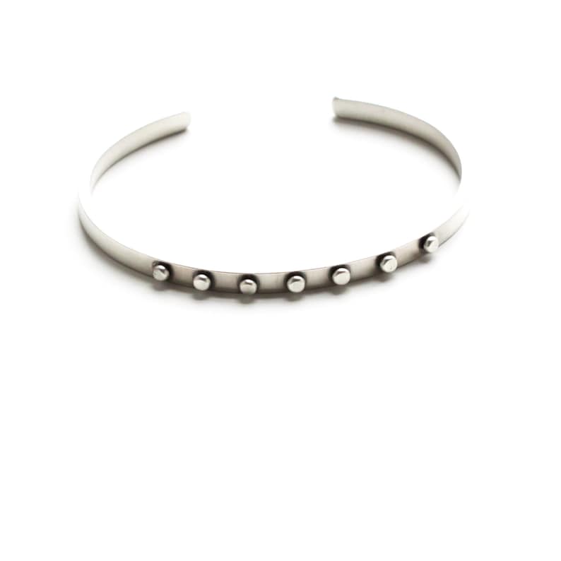 May include: A silver-colored cuff bracelet with a row of small, round, silver studs. The bracelet has an open design and is set against a white background. The jewelry is simple and modern.