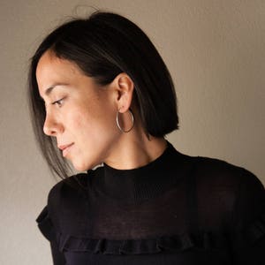 Puede incluir: Una persona con un jersey negro de cuello alto con un detalle de volantes. Lleva pendientes de aro plateados. La persona tiene el pelo oscuro y mira hacia abajo.