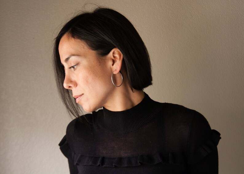 Puede incluir: Una persona con un jersey negro de cuello alto con un detalle de volantes. Lleva pendientes de aro plateados. La persona tiene el pelo oscuro y mira hacia abajo.